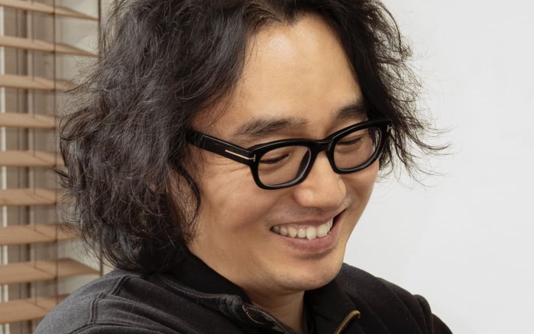 Juyong looks down and smiles. He is wearing glasses and a black polo shirt.