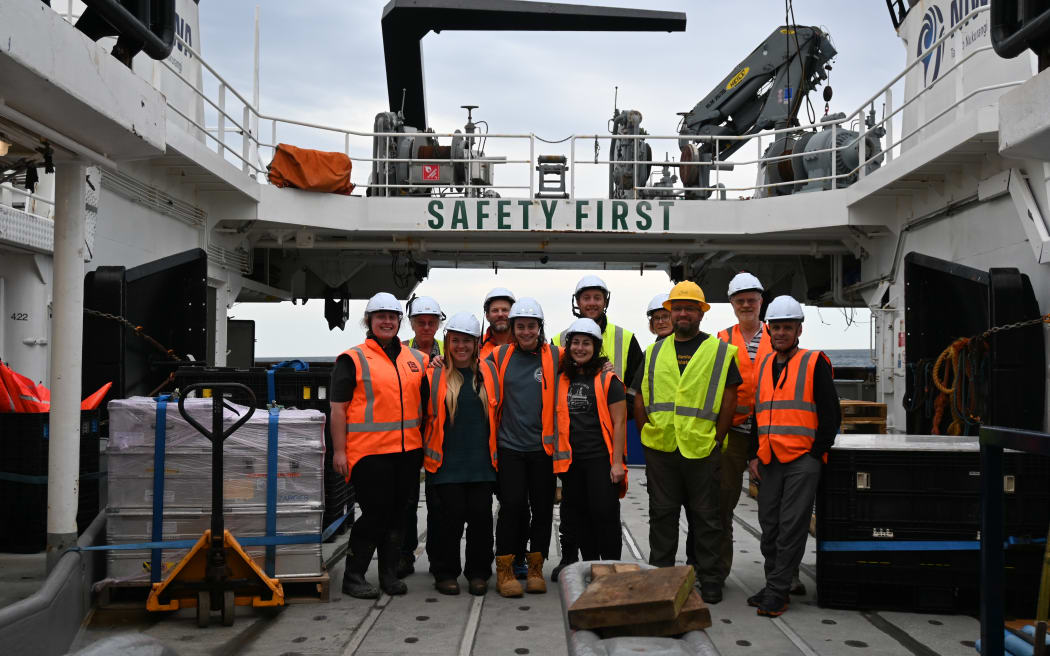 11 people in hard hats and high vis vests stand on the deck of a ship for a group photo. There is equipment on either side, and a bridge above that has 'Safety first' written on it.