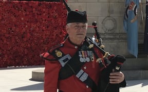 Les Kenfield is standing in front of a war memorial. He is in a kilt and red military jacket. He is holding his bagpipes.