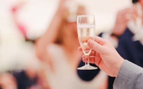 Glass of champagne being held up in celebration iwth partygoers in background.