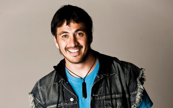 Tammy Davis is a young smiling man wearing a denim vest, blue t-shirt and pounamu pendsnt.