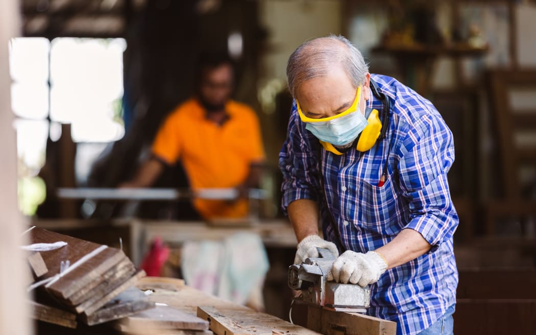 asian senior man carpenter wear protective mask dust and headphone working with electric planer In his own woodworks.