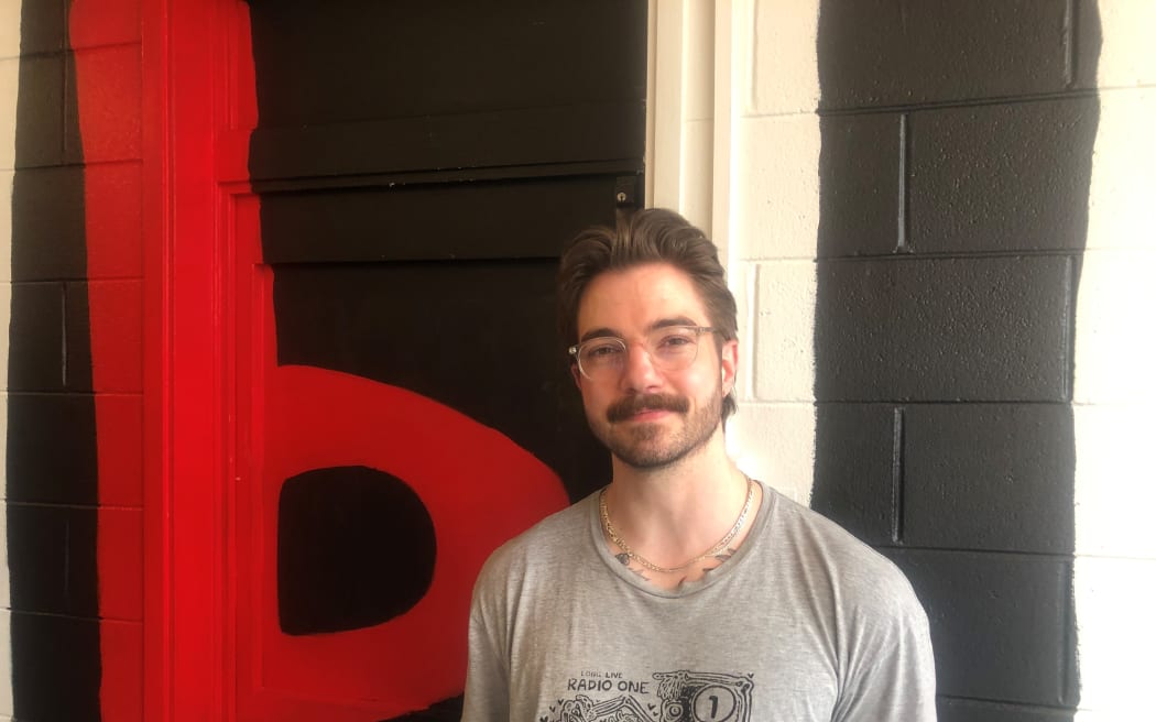 Tom smiles at the camera. He stands in front of a black wall with a large "95bFM" logo. He is wearing a grey tshirt with "LONG LIVE RADIO ONE" written on it in black, with a cartoon of a boombox.