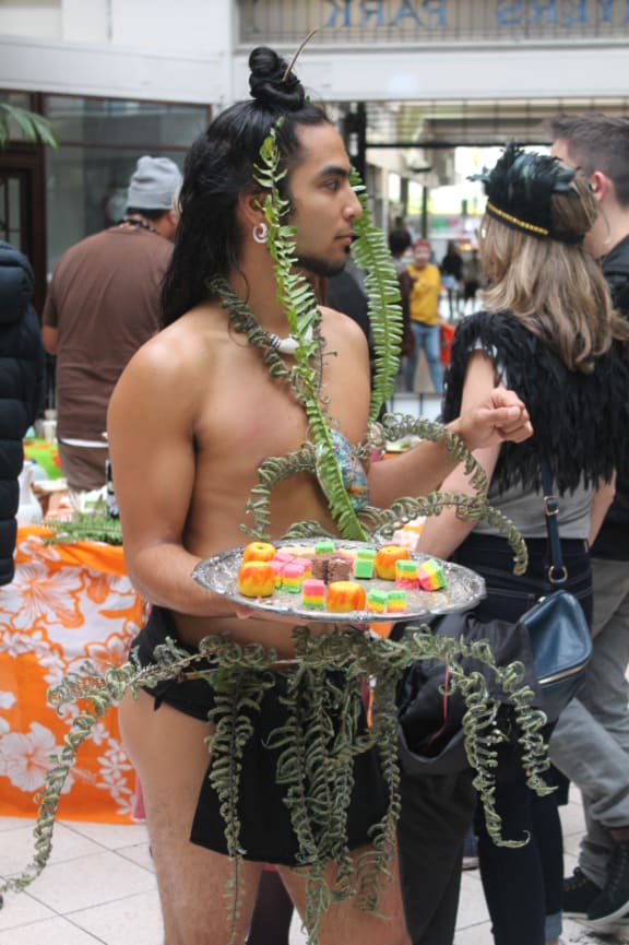 An image of dancer and performance poet Dåkot-ta Alcantara-Camacho  handing out small cakes while dressed in leaves.