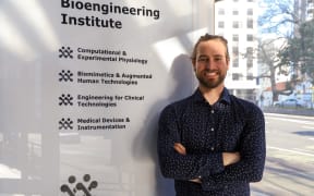 A man stands in front of a sign that says 'Bioengineering Institute'. He is smiling, wearing a navy shirt, and has his arms folded across his chest.