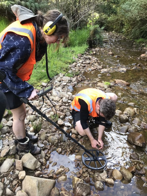 Nadine Cooper & James Scott search the riverbed to find what the metal detector has picked up. James is scooping stones out of the river bed while Nadine stands beside him holding the metal detector.
