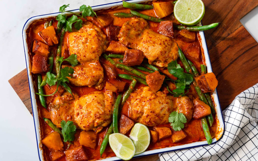 A rectangular oven tray containing red curry chicken and pumpkin tray bake, with green beans, coriander leaves and lime wedges.