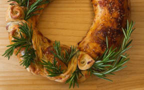 A Caramelised Onion Sausage Roll Wreath on a wooden board, decorated with sprigs of fresh rosemary.