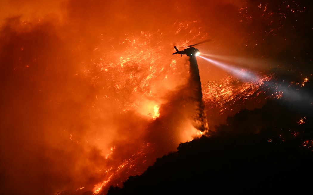 A fire fighting helicopter drops water as the Palisades fire grows near the Mandeville Canyon neighborhood and Encino, California, on 11 January 2025. The Palisades Fire, the largest of the Los Angeles fires, spread toward previously untouched neighborhoods January 11, forcing new evacuations and dimming hopes that the disaster was coming under control. Across the city, at least 11 people have died as multiple fires have ripped through residential areas since January 7, razing thousands of homes in destruction that US President Joe Biden likened to a 