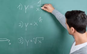 21205086 - male teacher with his back to the camera writing a mathematical formula on the blackboard in a classroom while teaching a class