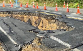 A photo of damage to the state highway shows cracking and slumping in the road with road cones around it.