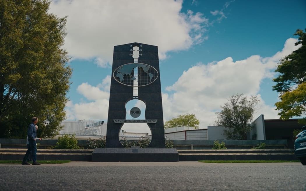 A large monument on the side of a road in Gore. It shows a silhouette of a guitar with a flat image of the globe on top of the frets. People stand to the side, looking at it.