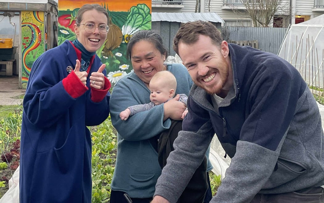 This is a photo of Wilby LeHeux signing the lease agreement for the new community garden space. He is stood outside in their existing garden. Alongside him are Janel Lou Yee Mei. Janey is also holding a baby. Sophie Bond is next to her giving a thumbs up to the camera.