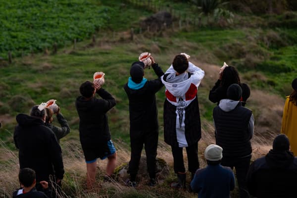 A group of people stand outside facing away from the camera, some holding pūtātara (a conch shell trumpet) which they appear to be blowing