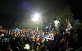 Thousands of people huddle around a small stage at Ihumātao enjoying a free concert.