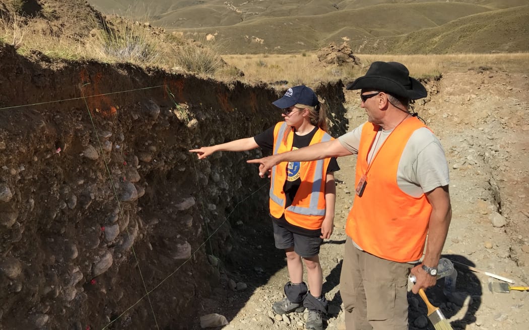 Mark and Ashleigh are pointing to the wall of the trench which has been marked out with string and coloured plastic tags. Mark holds a paint brush and a bottle of water.