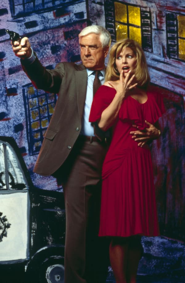 Leslie Nielsen, a white-haired man in a brown suit, aims a gun and clutches Priscilla Presley - a blonde-haired woman in a burgundy dress.