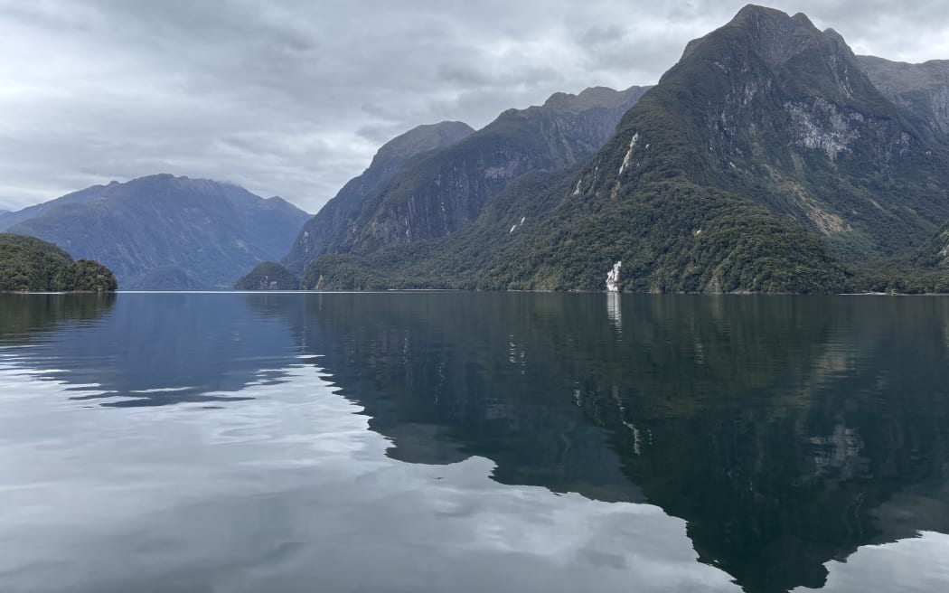 A landscape shot of a fiord with still dark water, reflecting steep-sided bushclas mountains with waterfalls plunging down them. The sky is grey and cloudy.