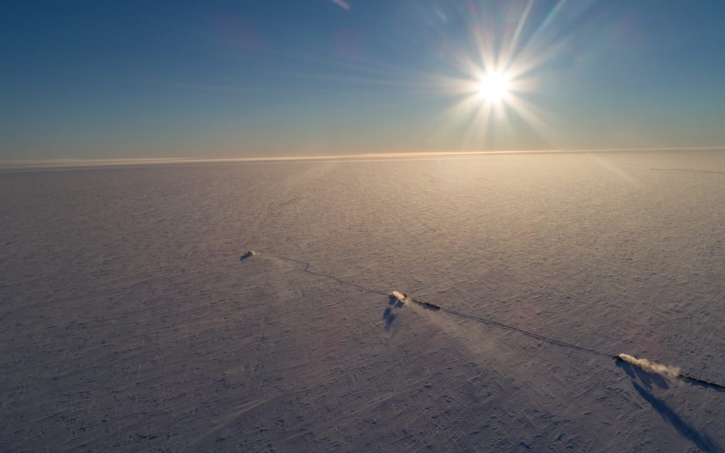 An aerial image looking down on the flat white expanse of an ice shelf, beneath a white sun and darkening indigo sky. Three vehicles, that look small from this height and angle, are traversing the expanse from left to right in a diagonal line.