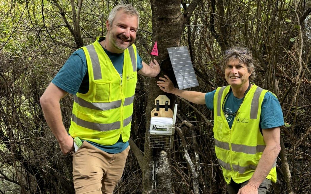 Two men in blue t-shirts and fluoro yellow hi-vis vests stand next to a smart trap attached to a tree.