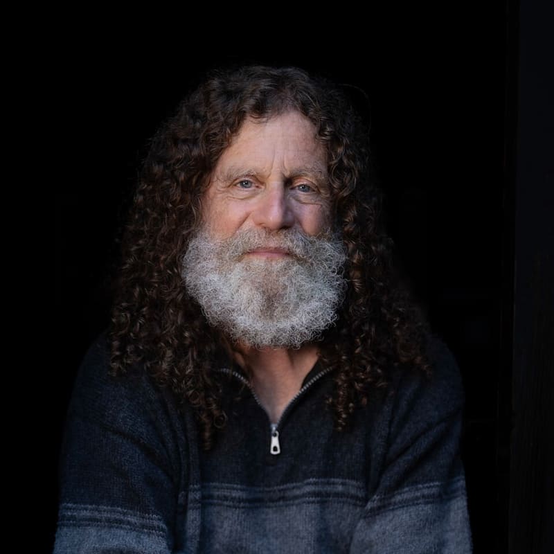 Robert Sapolsky has long brown hair and a white beard and sits on a stool holding what appears to be a human skull.