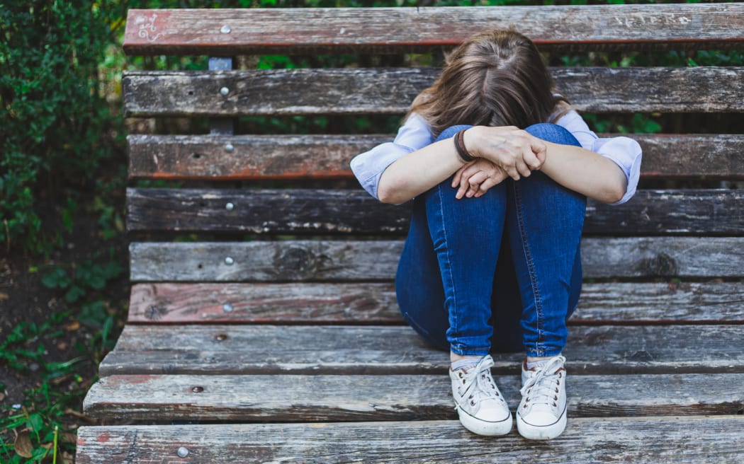 Sad and depressed girl sitting alone on a bench in the park - Young woman with brown hair having anxiety problems outdoors - Stressed and frustrated female in need for help.