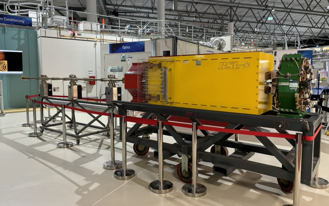 A big yellow metal box with a complex green wheel on one end, and a red contraption on the other, sits on a bench in a warehouse with a high ceiling.