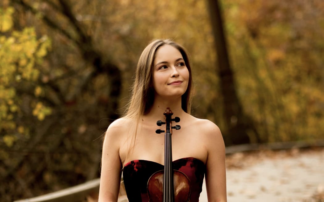 Violinist Geneva Lewis sits cross-legged with her violin held upright in her lap.