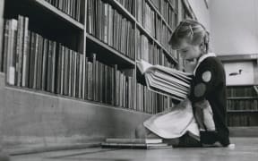 Young girl reading book in front of set of full library shelves