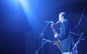 Saxophonist playing into a microphone, suffused in blue light.