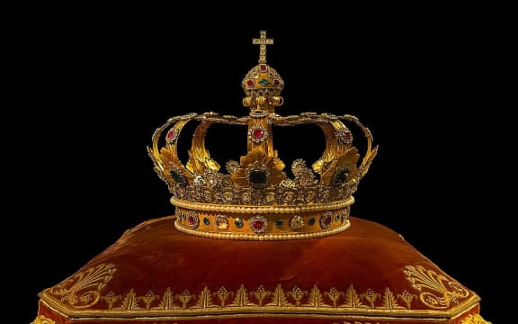 A gold crown with rubies and emeralds, sitting on a red velvet cushion