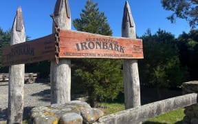 The entryway to Re-Engineered Ironbark, showing four large upright wooden poles capped with steel with a large wooden sign. In the foreground, a gateway made of riverstones