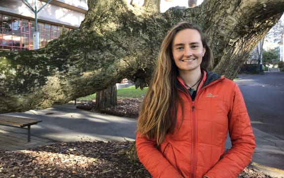 Edin is standing in front of a tree, smiling, wearing a red jacket.