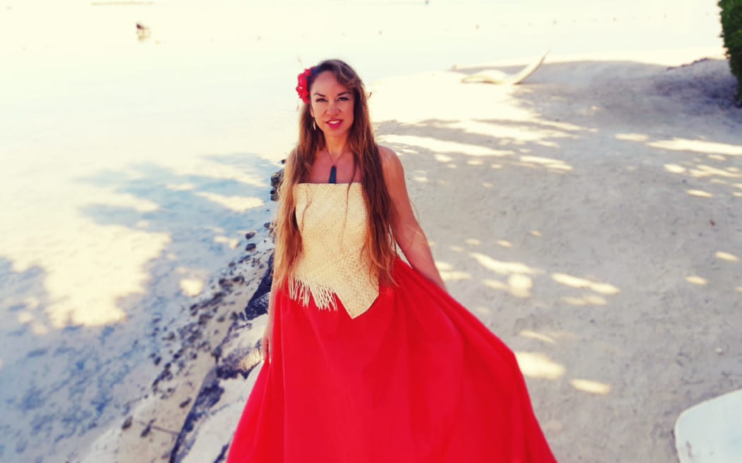 Toni Huata stands on a white sand beach, wearing pounamu and a red dress with a red frangipani in her hair.