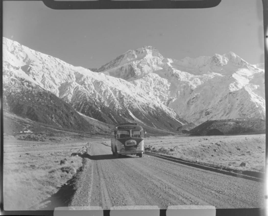 Mount Cook and Southern Lakes Tourist Coach on the Hermitage Road, Mount Cook Village, with Mount Sefton beyond, Mount Cook National Park, Canterbury Region. Whites Aviation Ltd: Photographs. Ref: WA-41978-F. Alexander Turnbull Library, Wellington, New Zealand. /records/30656529