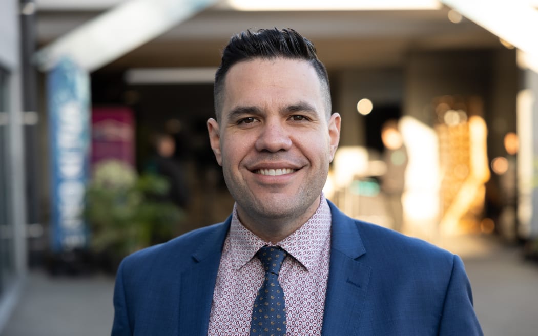 Dr Jeremy Tātere MacLeod is standing in front of a building. The background is in very soft focus so it's impossible to tell what it is. Jeremy is in sharp focus. He is wearing a blue suit and tie with a patterned purple shirt.