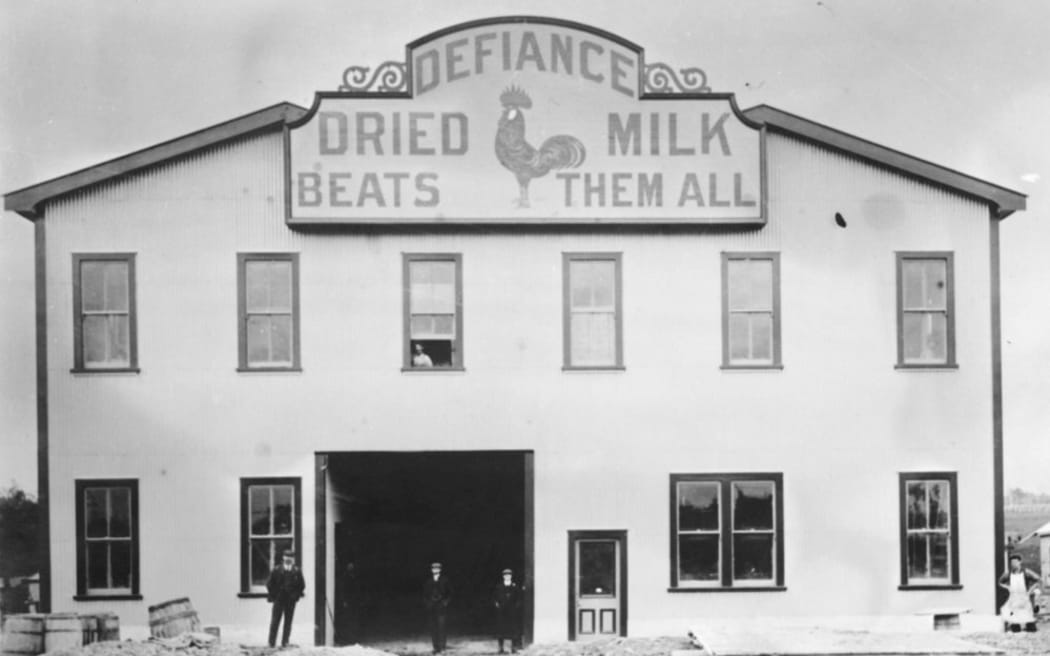 A historic photograph showing the Defiance milk powder factory front. The sign says "DEFIANCE - DRIED MILK - BEATS THEM ALL". The company's logo, a rooster, sits in the middle of the sign. Several men pose for the photo in front of the factory doors.
