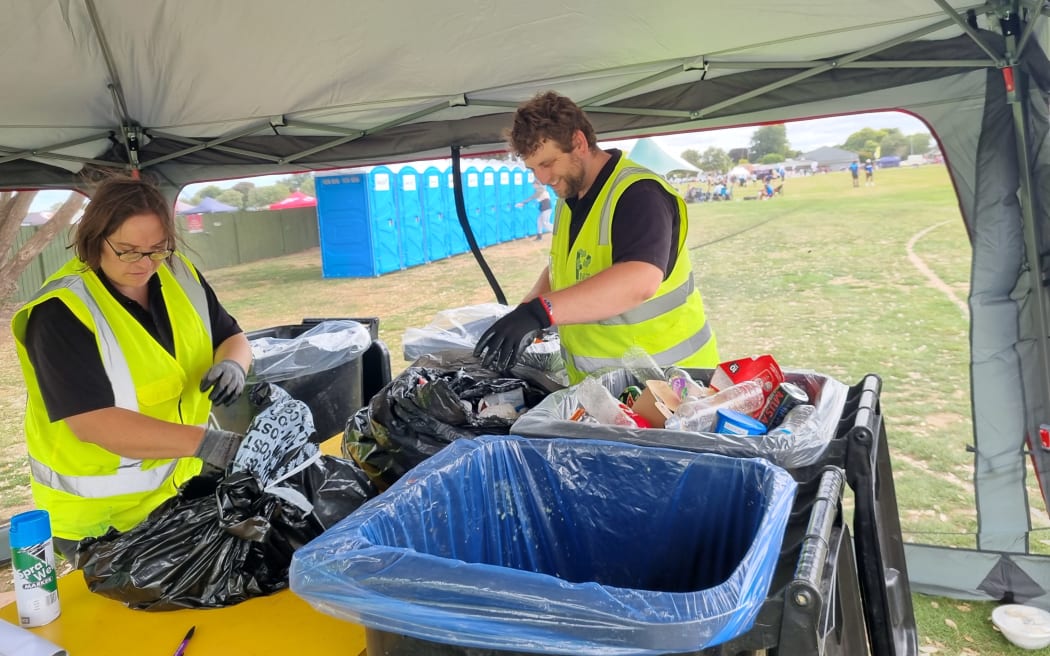 Two people in hi-vis jackets stand under a gazebo sorting through rubbish bags by hand.