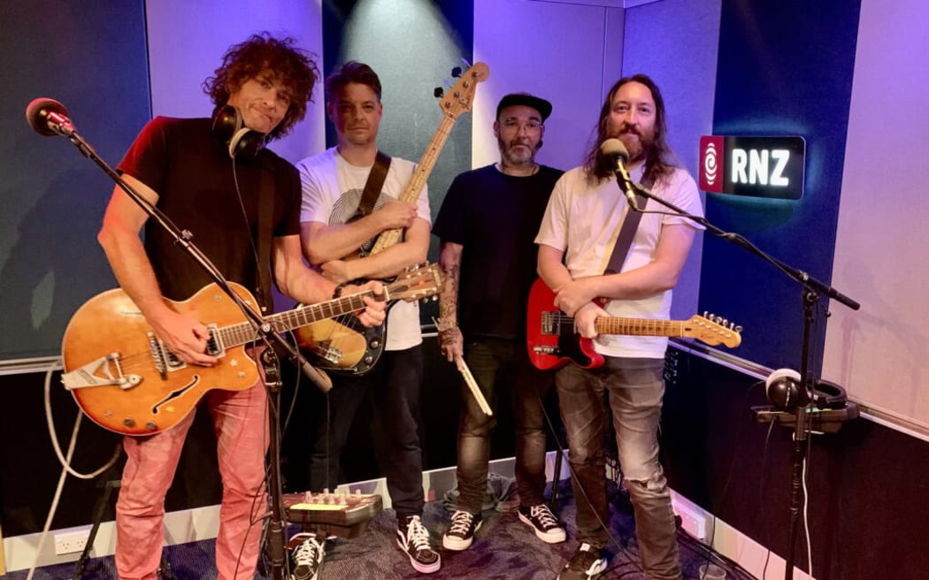 The four members of Voom with their respective instruments against a teal and white sound-paneled wall. They are lit with a purple light from behind them.