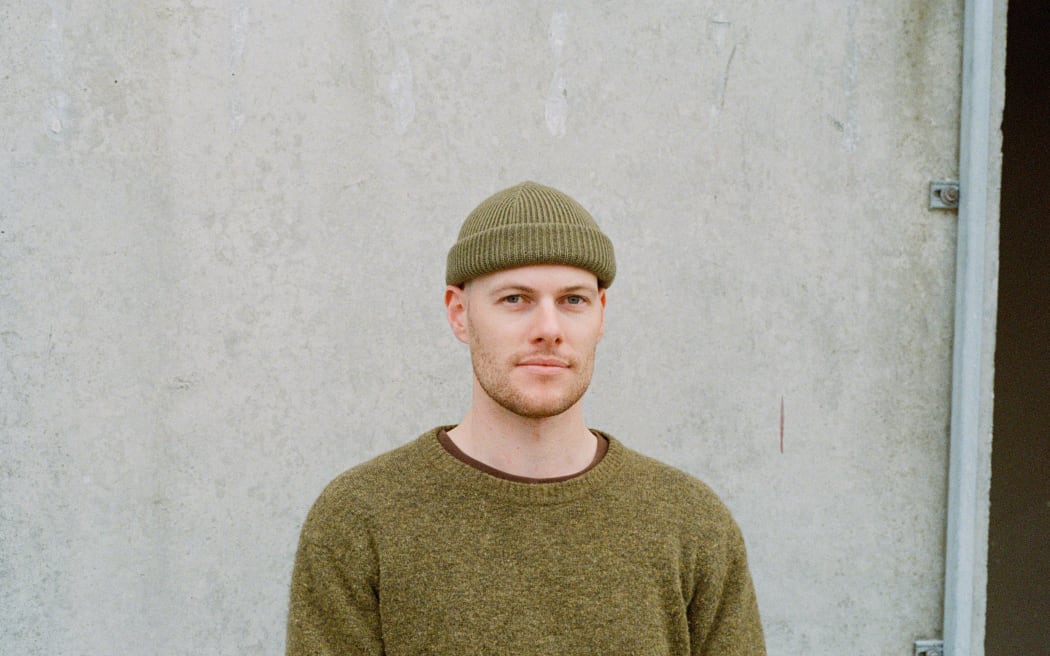A man in a green beanie and top looks at the camera. He stands in front of a white wall.