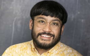 David smiles at the camera. He wears a yellow patterned shirt and gold-rimmed glasses.