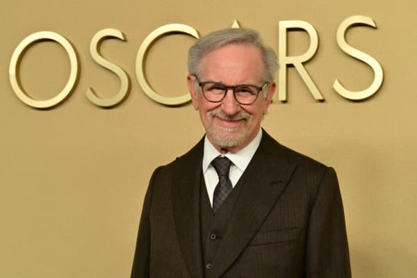 Steven Spielberg in dark-rimmed glasses and a dark suit, smiles against a gold wall that reads 'Oscars'.