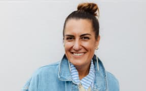 Qiane Matata-Sipu is a smiling woman with her hair pulled back, long white earrings and a pale denim jacket.