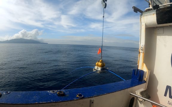 A view of Whakaari / White Island from the deck of a ship. In the foreground a yellow sensor being lowered into the ocean from a crane.