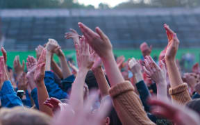 Audience with hands raised at a music festival and lights streaming down from above the stage.