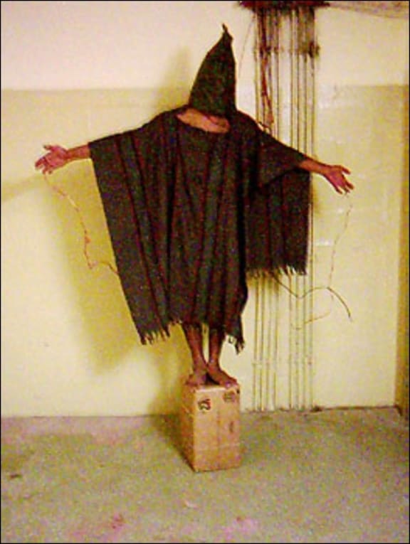 Detainee with bag over head, standing on box with dummy wires attached, under threat that stepping down from the box would cause his electrocution. Abu Ghraib 2003.
