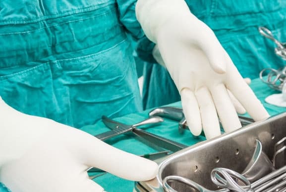 A surgeon in scrubs and white gloves with a tray of instruments.
