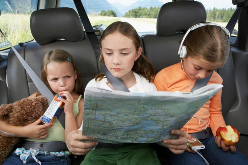 Three children in the back seat of a car, one drinking juice, another reading a print map, and the third with headphones and an apple.