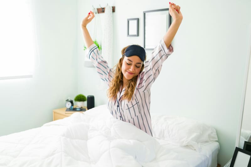 A woman wearing pyjamas and an eye mask gets up from bed stretching.
