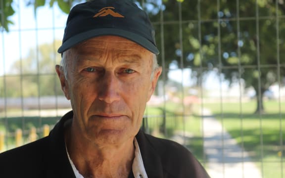 A man wearing a black polo shirt and a black cap with a mountain on it looks at the camera intently. Behind him in low-focus is a fenced-off, grassy area.